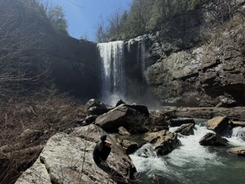 A hiker sitting on boulders below a tall waterfall